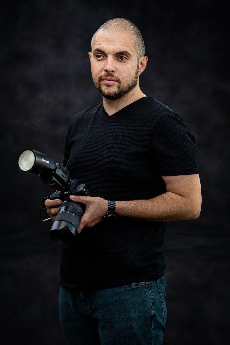 Atanas sostiene una cámara con flash, sobre un fondo oscuro de estudio
