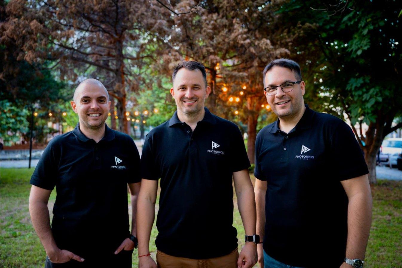Ilko, Rado y Atanas con los polos de PhotoBros, juntos en un parque al anochecer con luces de guirnalda detrás